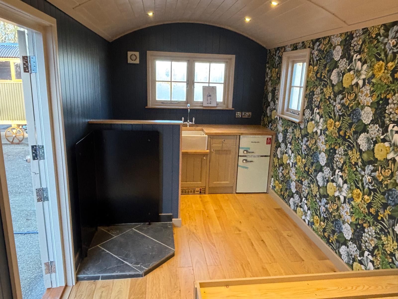 Small room with floral wallpaper, wood floor, black stove, and a compact kitchen nook by the window.
