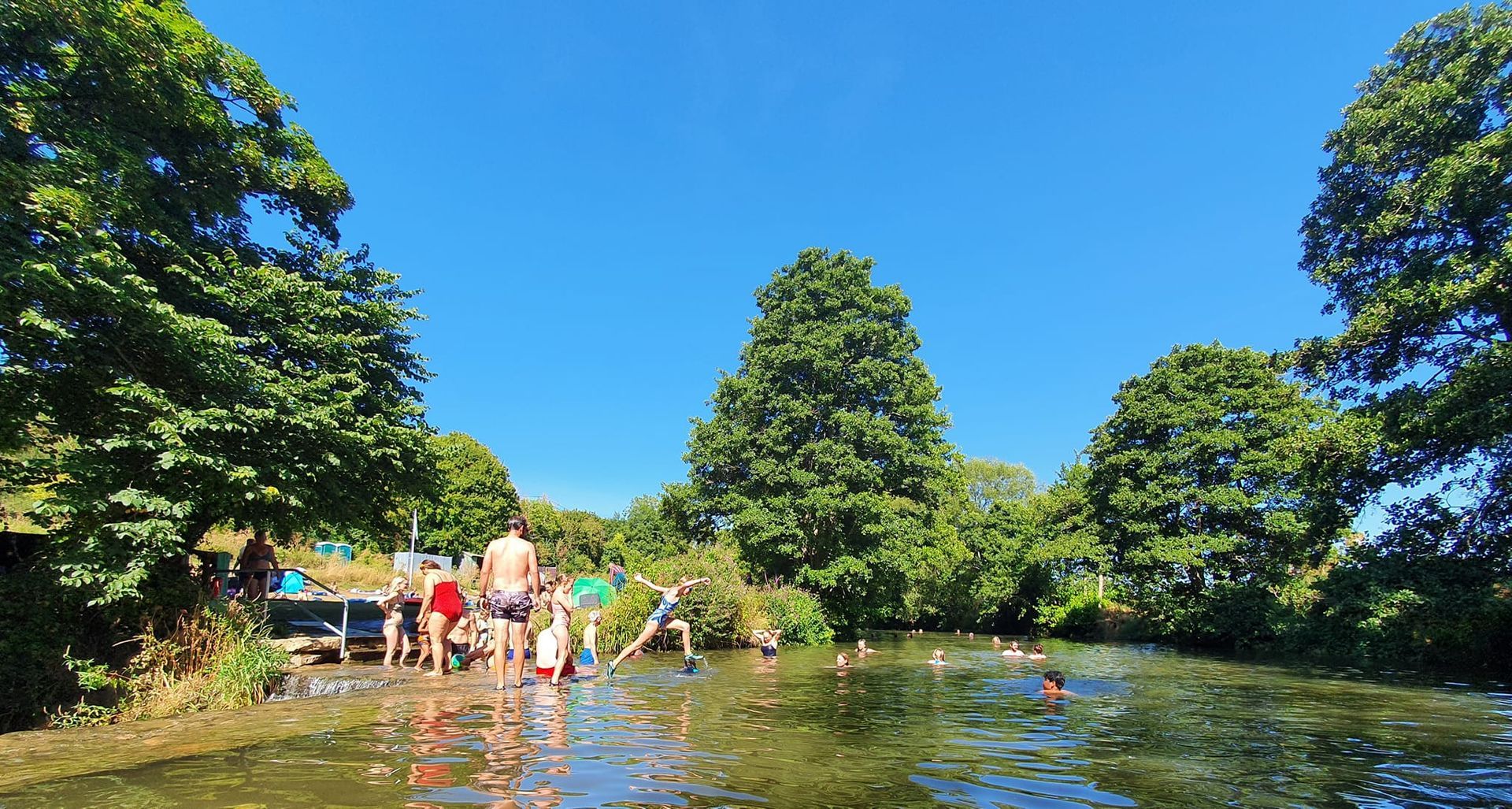 People wading and swimming in a shallow river under bright blue sky, surrounded by green trees.