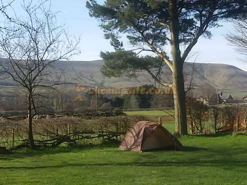 Small brown tent on grassy campsite with trees, stone walls, and hills in the background.