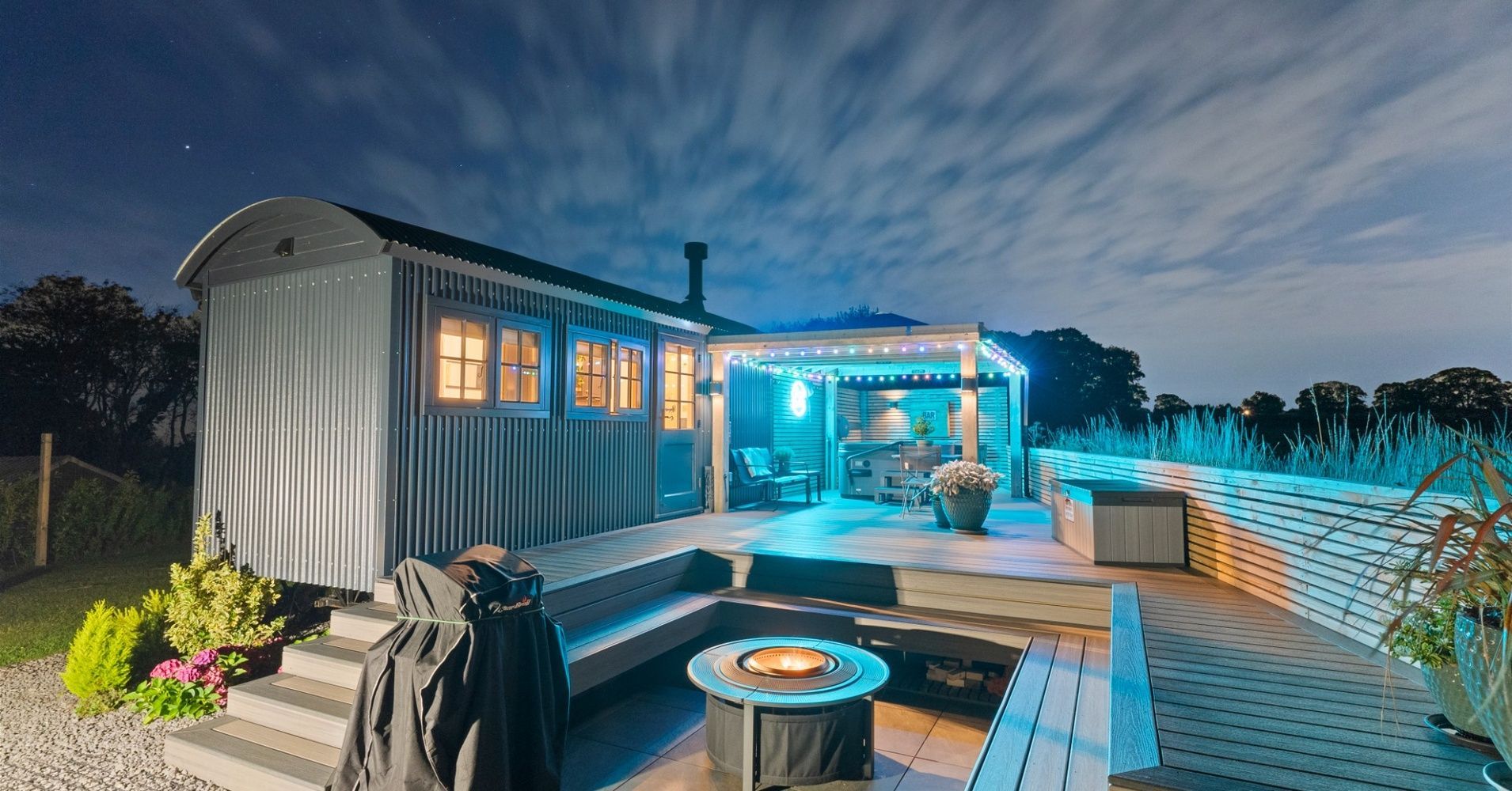 Cozy blue-lit tiny house with deck and hot tub at dusk, surrounded by snow.