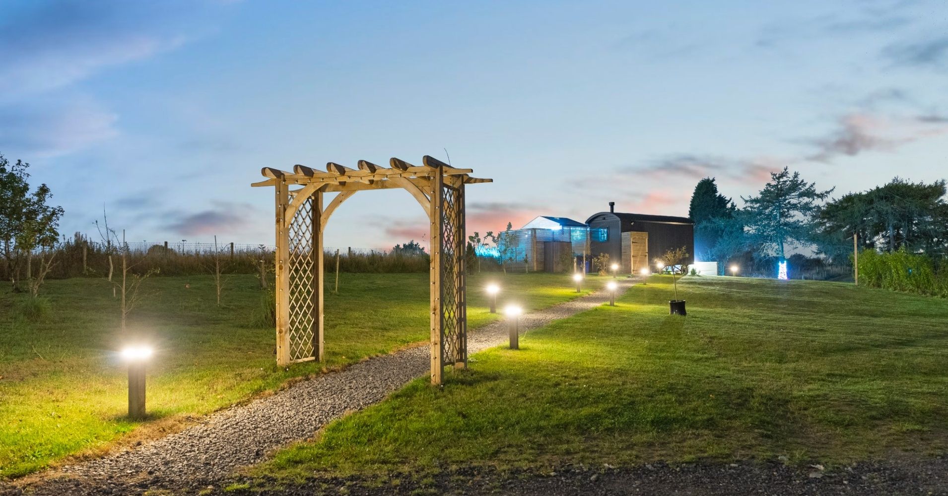 Lit garden path with a wooden arbor and distant buildings at dusk
