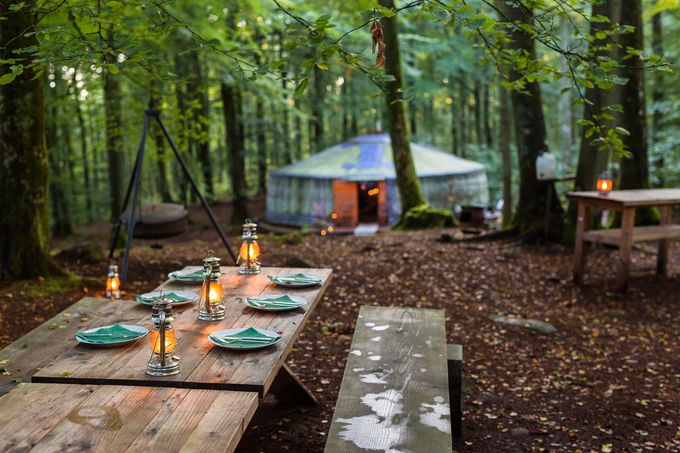 Lantern-lit picnic tables beside a green yurt in a wooded campsite at dusk