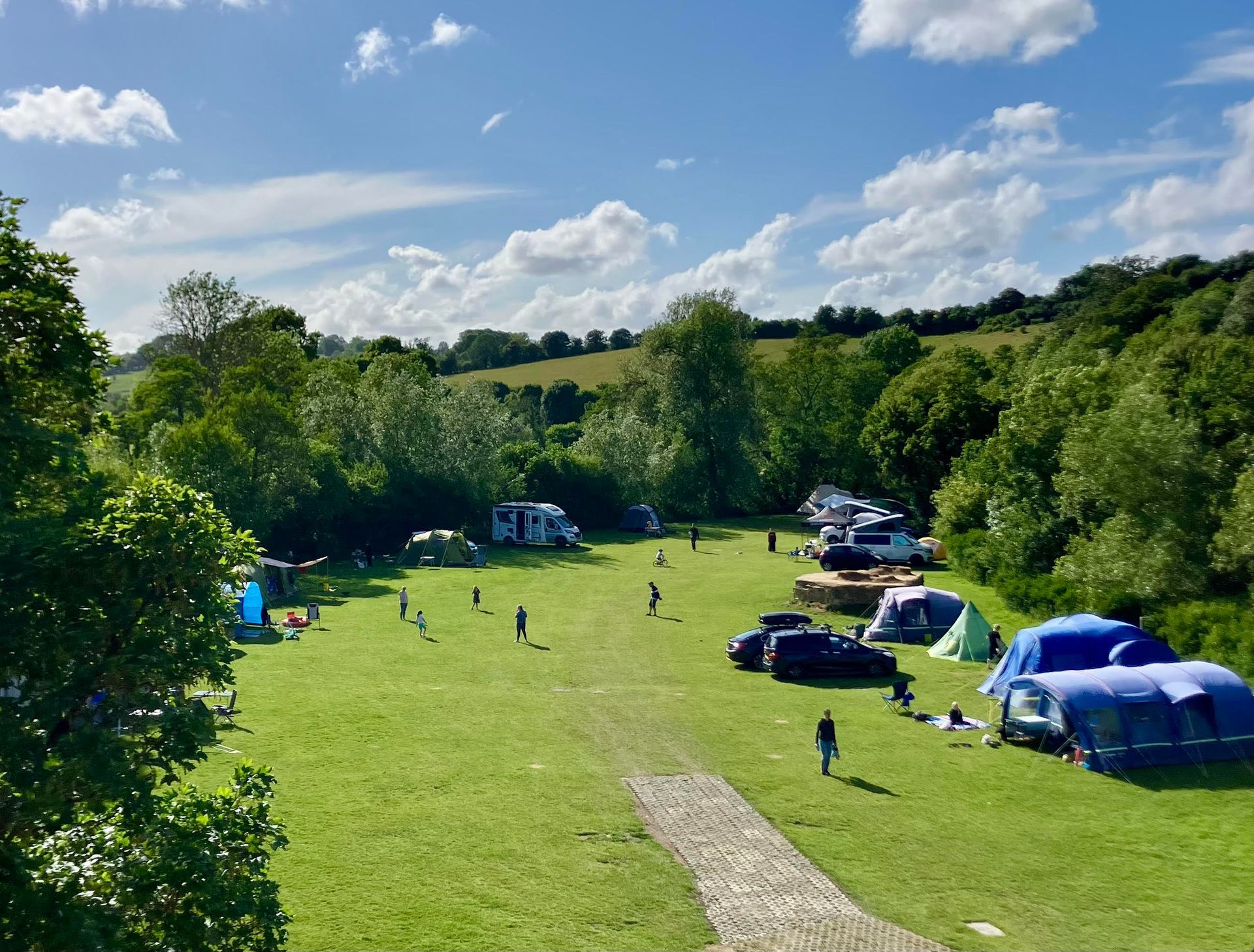 Sunny campsite on a grassy field with tents, parked cars, and people scattered among trees.