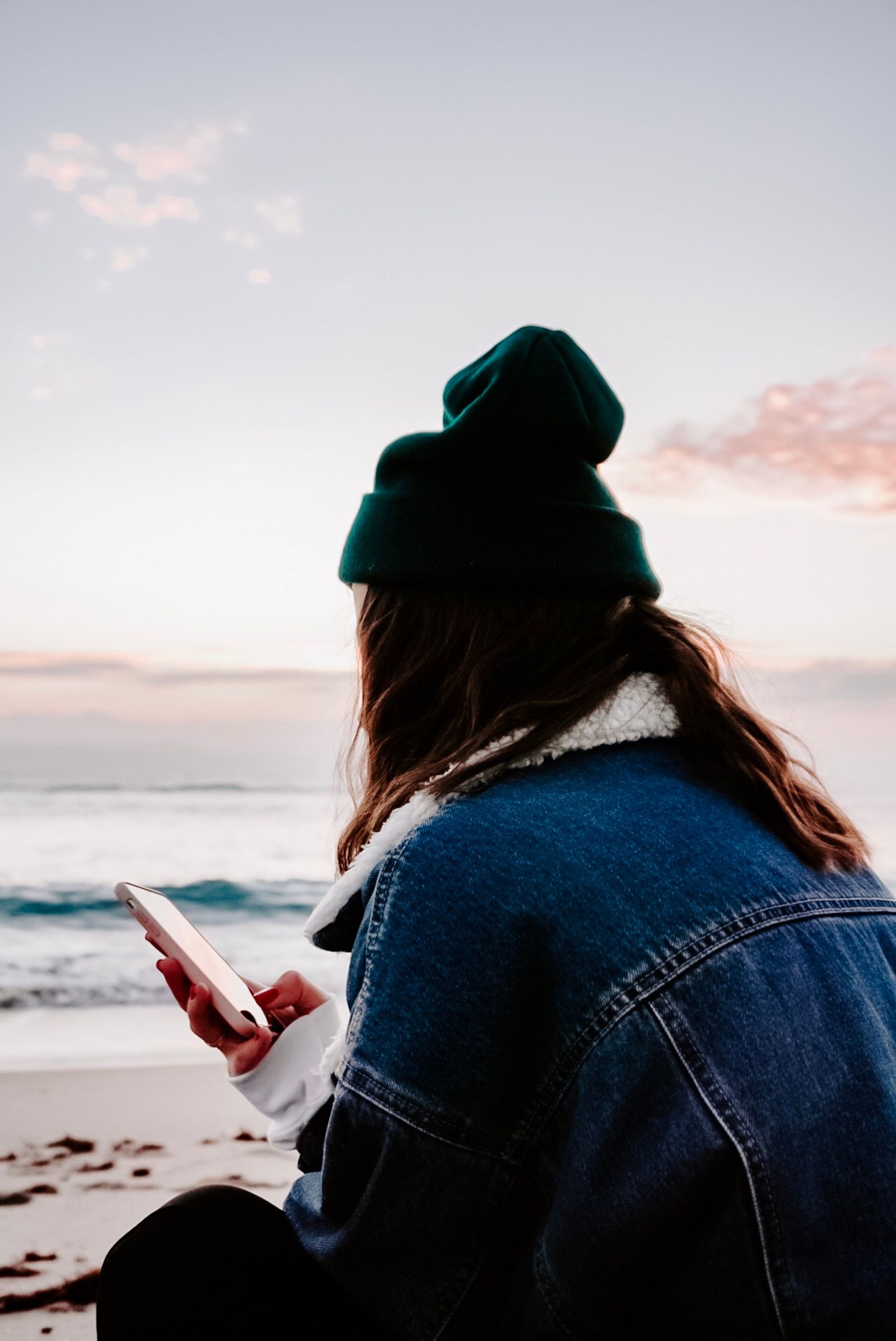 Person in a black beanie and denim jacket sitting by the shore at sunset, looking at a phone