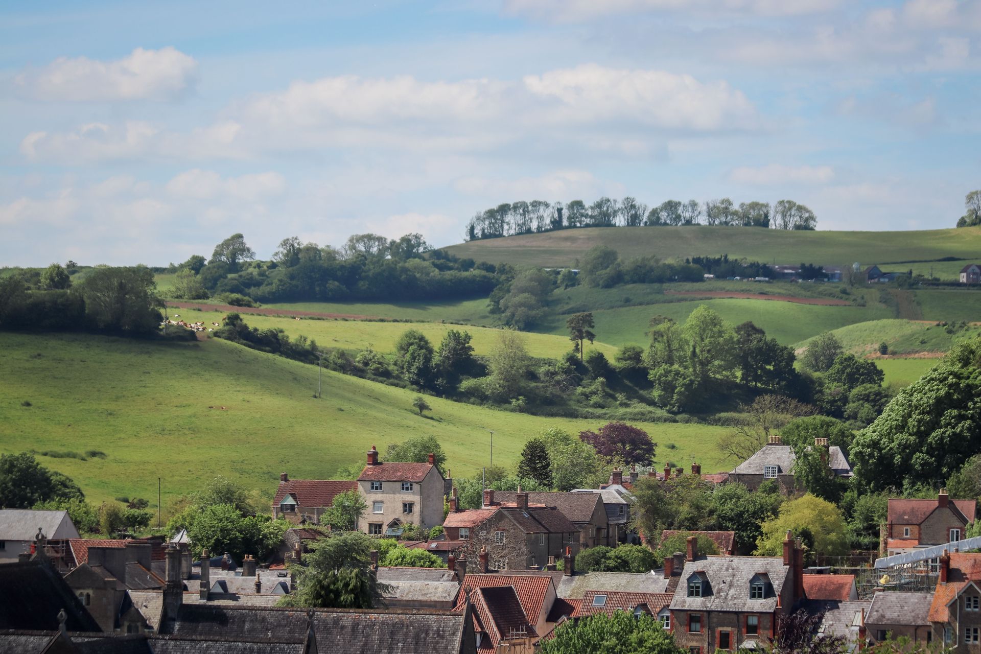 Green rolling hills above a village with red-roofed houses under a partly cloudy sky