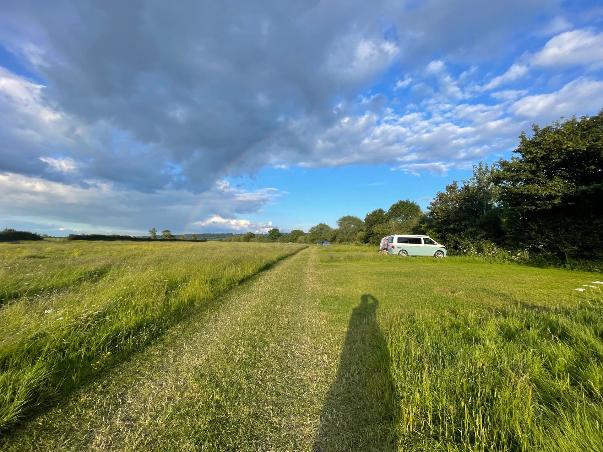 Grassy path through fields under a cloudy blue sky, with a white van parked by trees on the right.