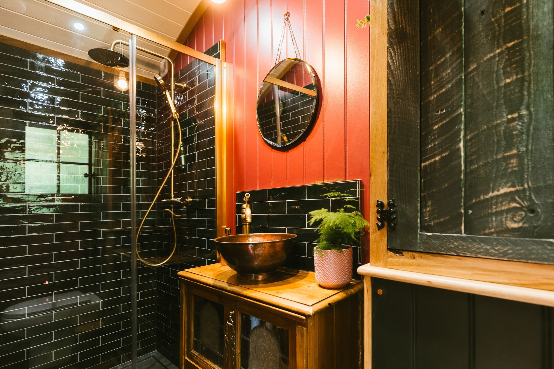 Modern bathroom with black tile walls, wood vanity, round mirror, and shower with glass door