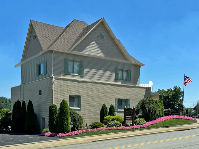 Large beige house with a steep roof, pink flowers, and an American flag under a clear blue sky