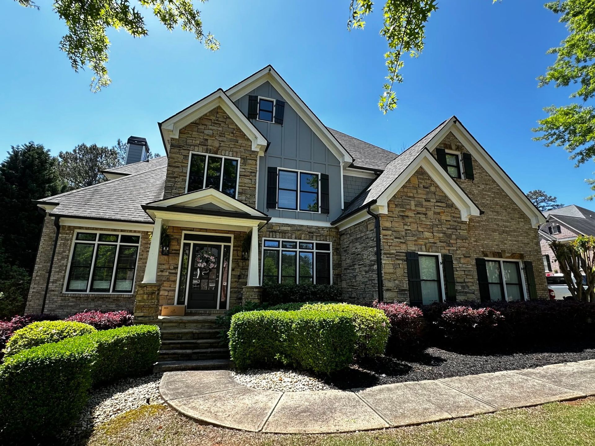Two-story stone house with gabled roof, front porch, and manicured shrubs under a blue sky