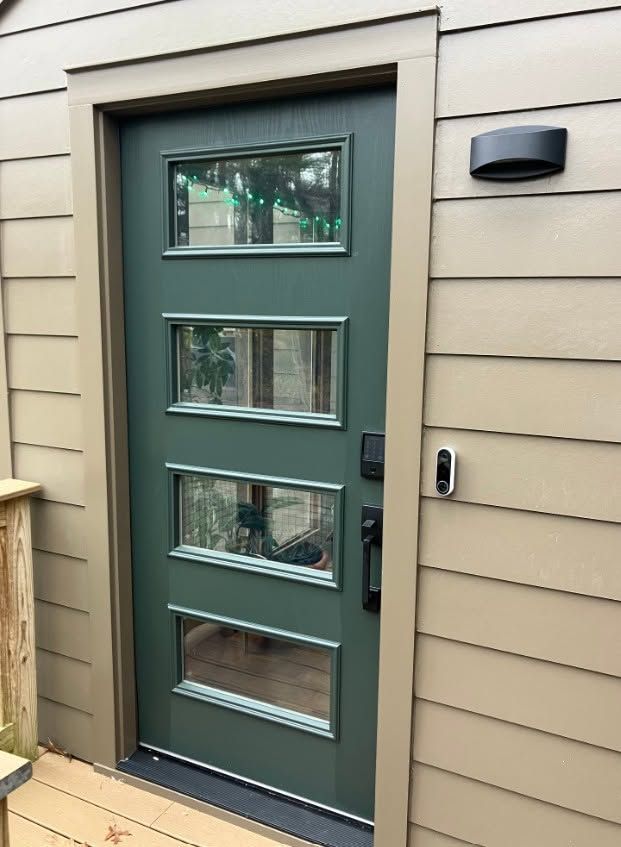Green exterior door with four small windows, set in beige siding with a wall light beside it