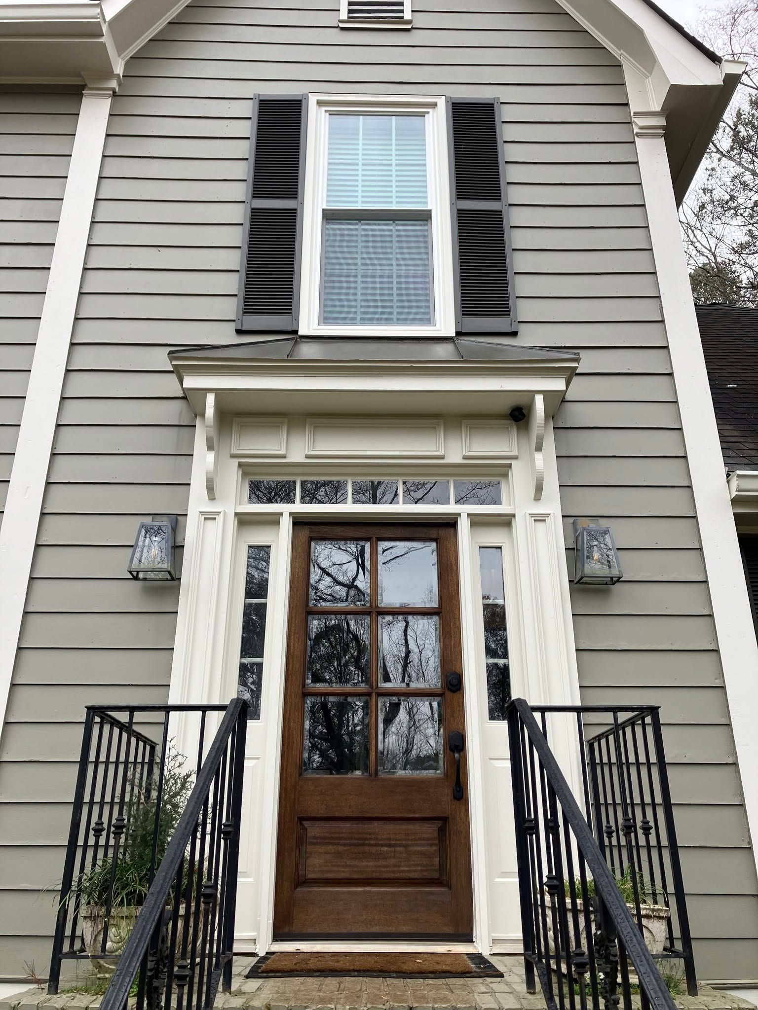 Front entrance of a gray house with wooden door, black shutters, and wrought-iron railings.