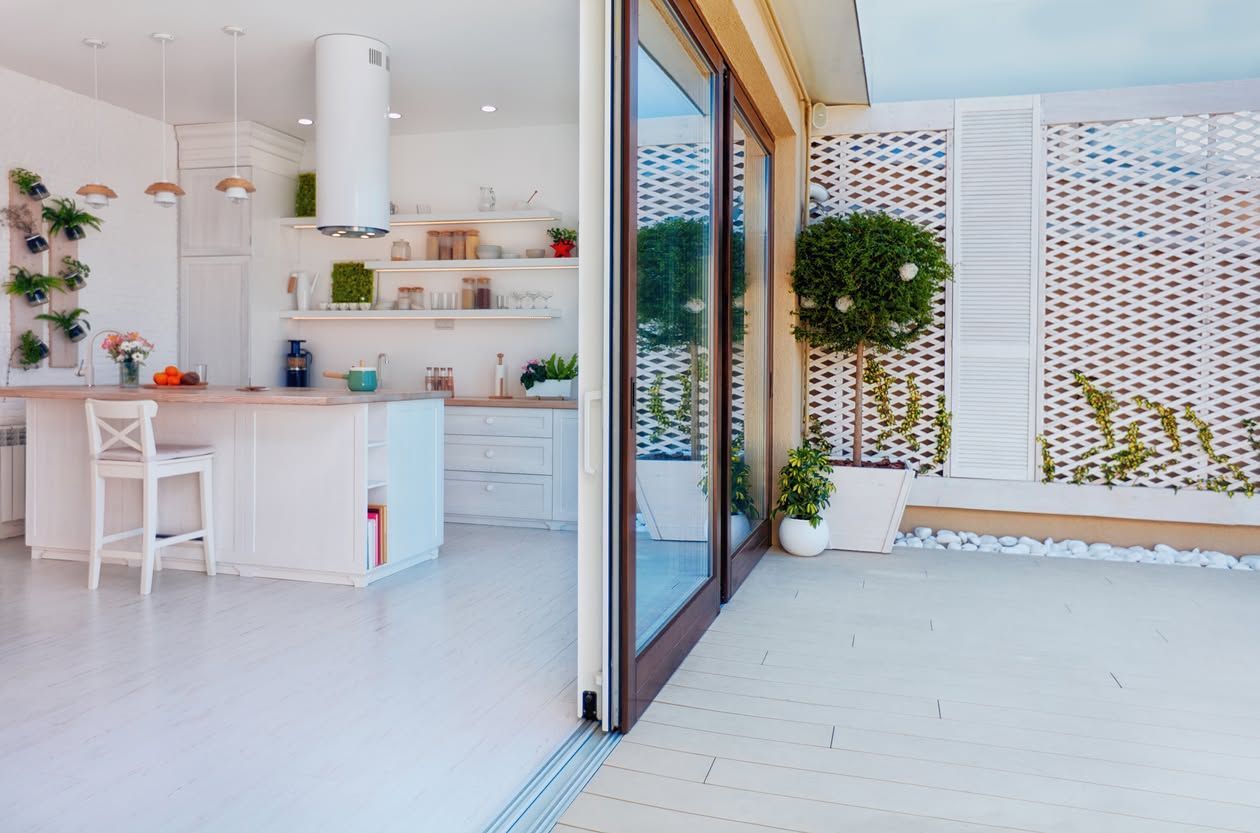 Bright white modern kitchen opening to a sunny patio with plants and woven privacy screen