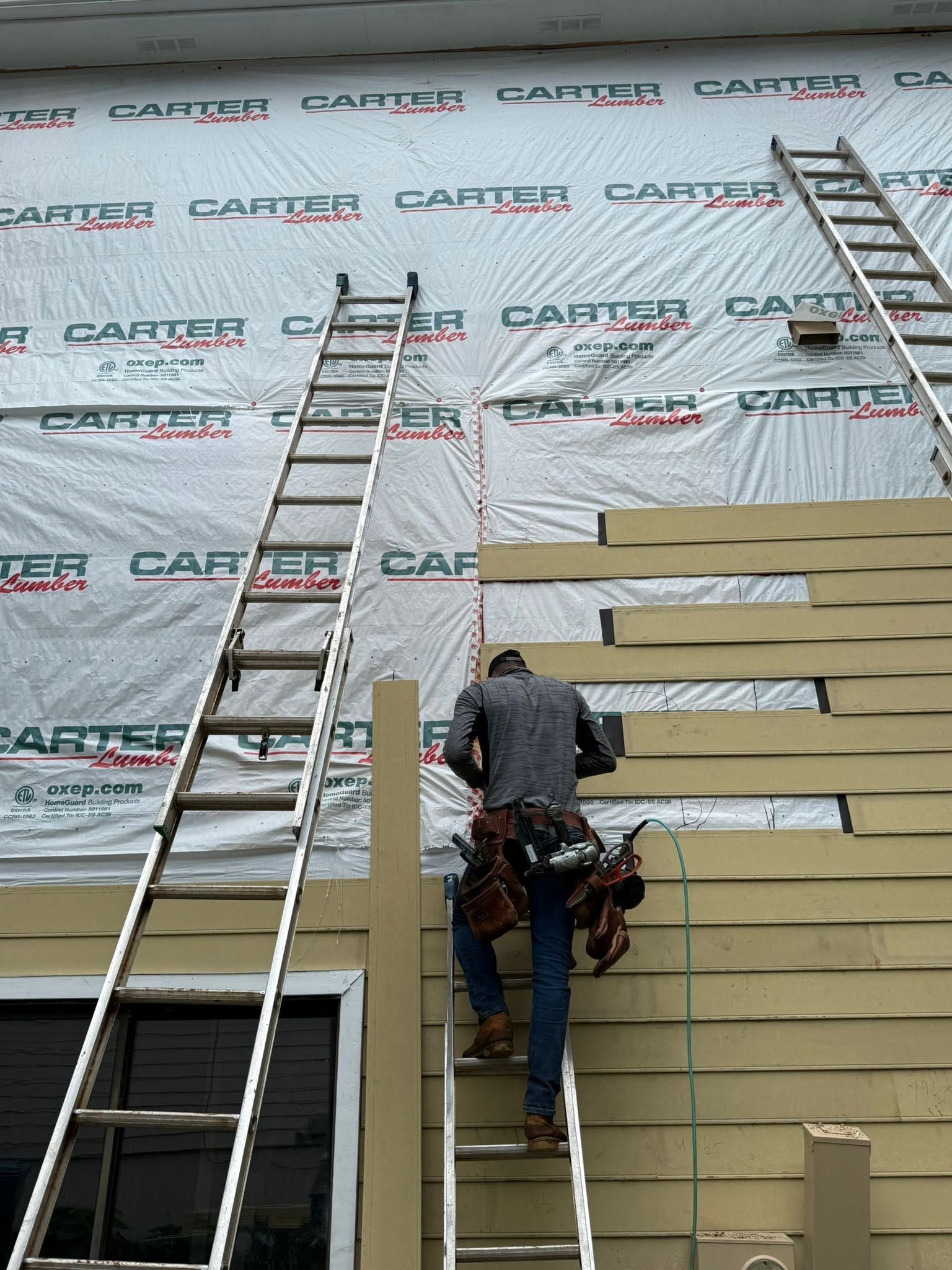 Worker on ladder installing beige siding on a building with wrapped upper wall and another ladder nearby