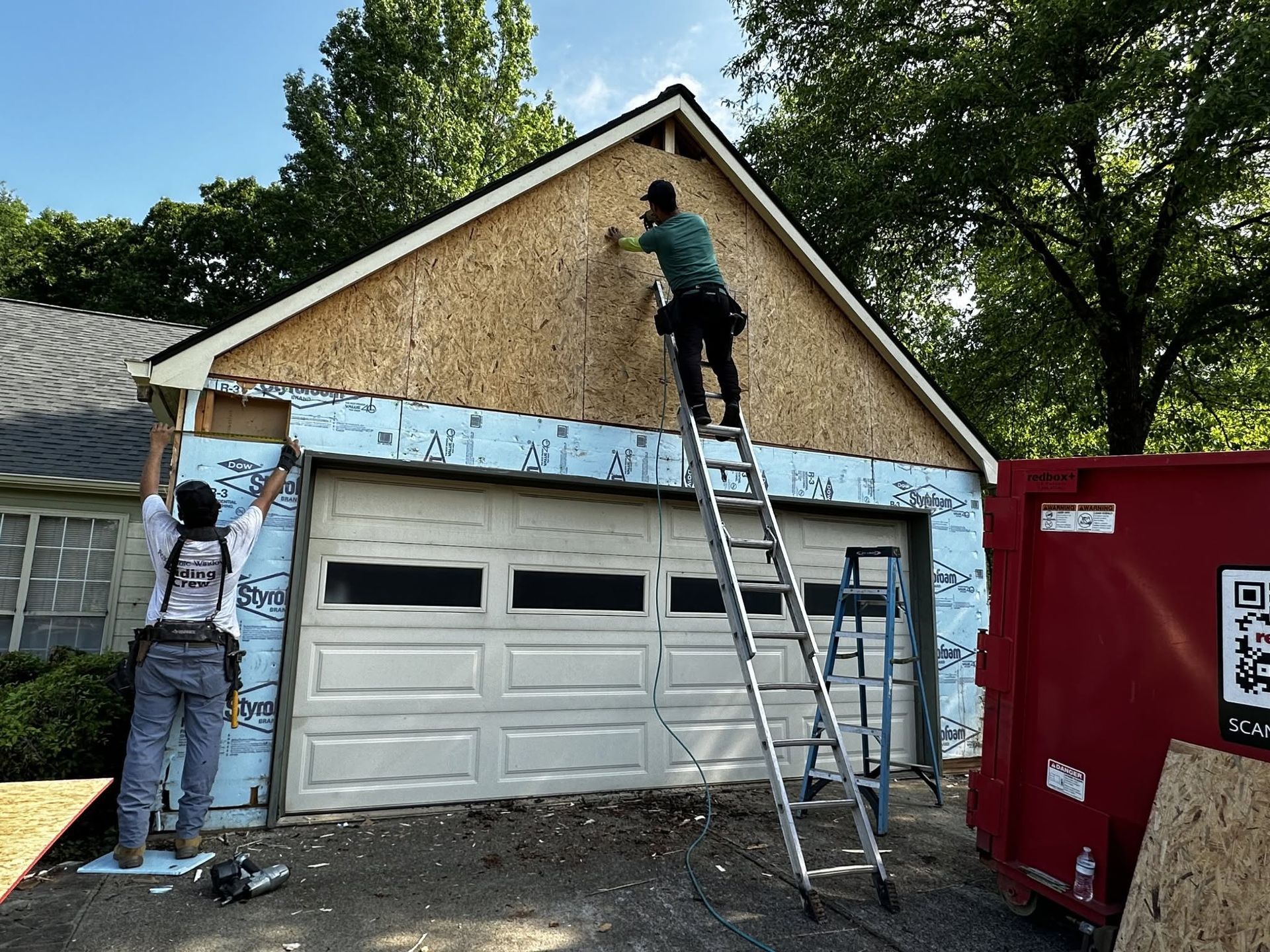 Two workers installing siding on a garage roof, with a ladder and red dumpster nearby.