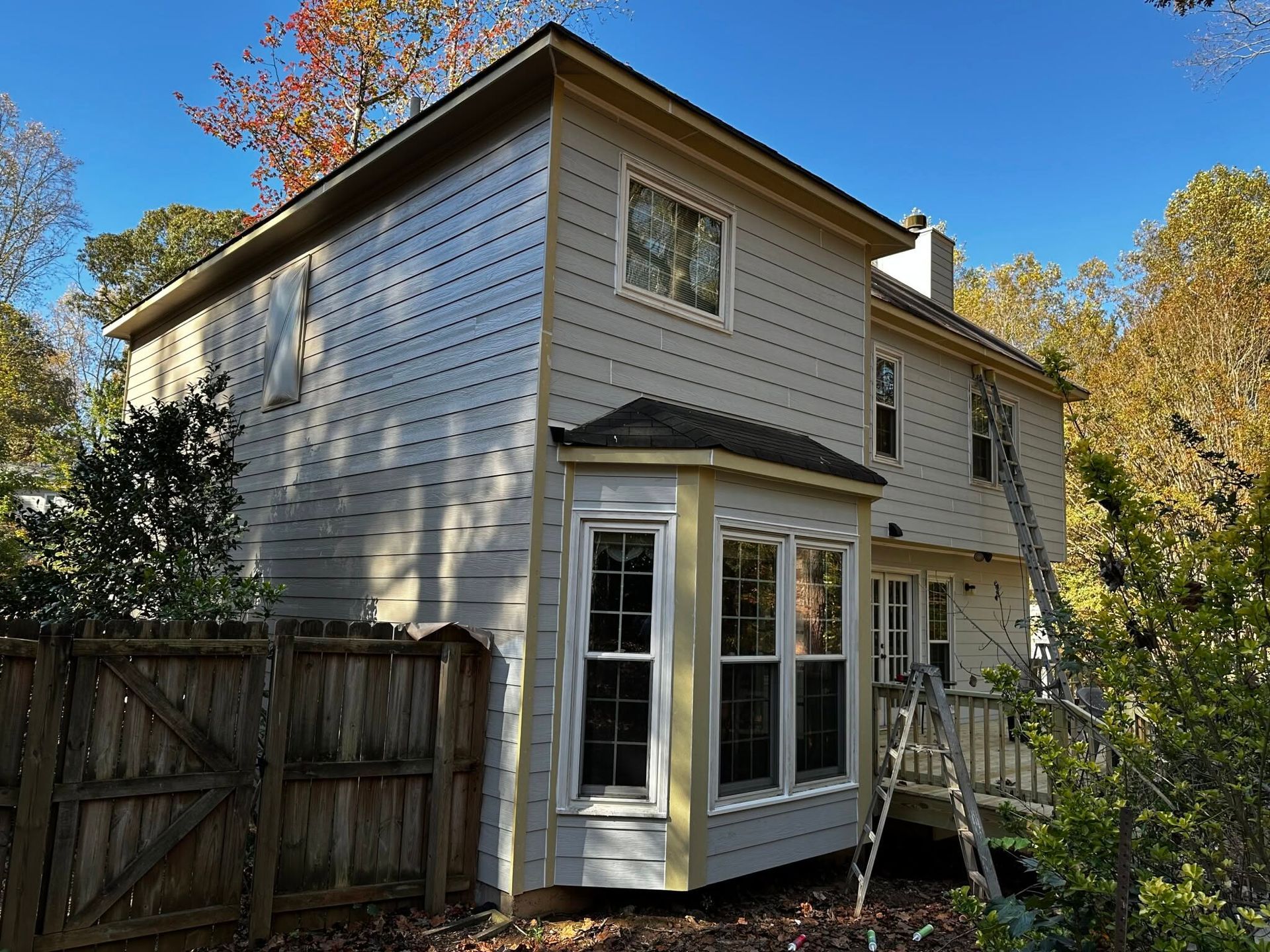 Two-story beige house with a bay window and fenced yard under a clear blue sky