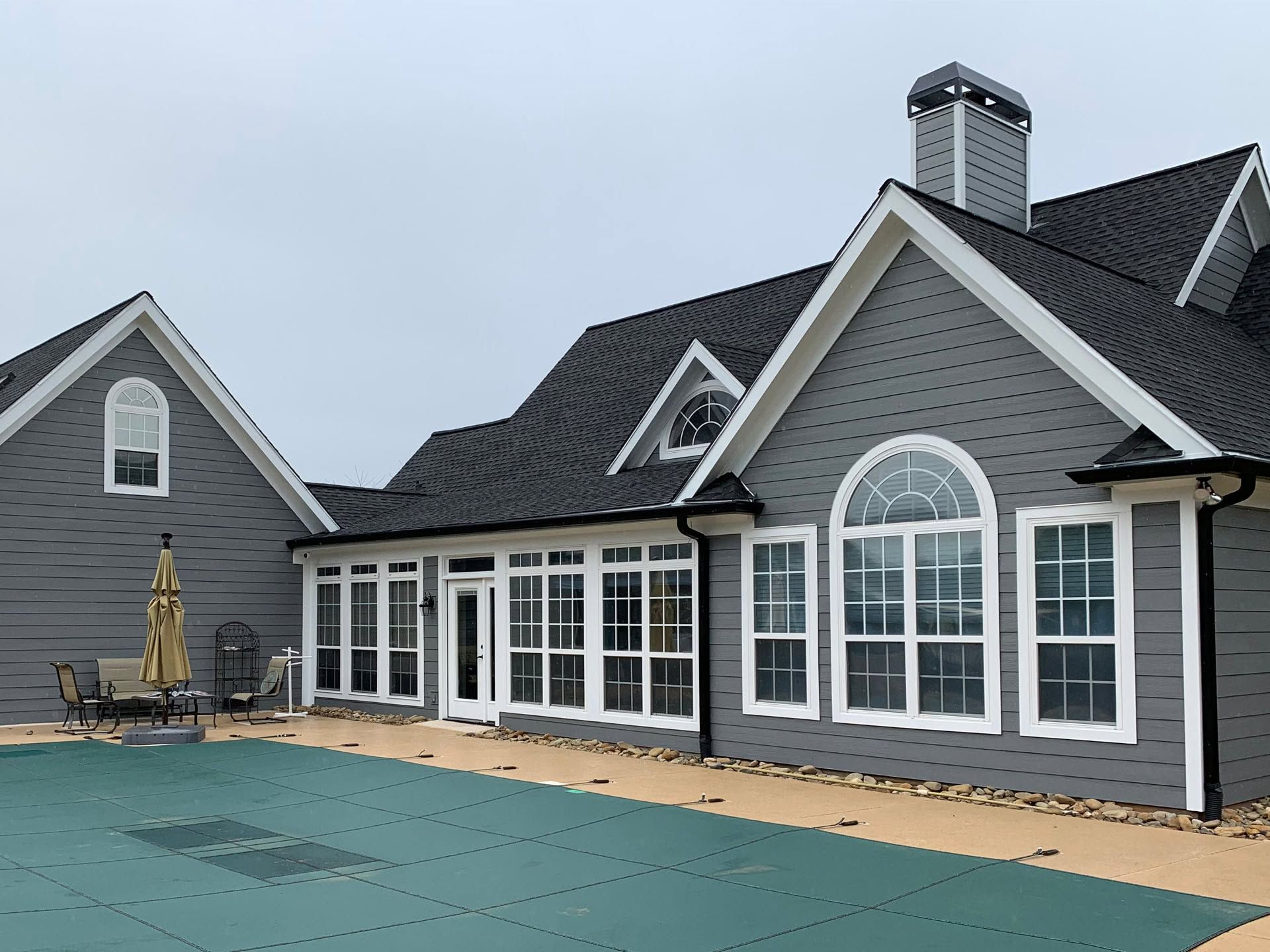 Large gray house with white-trimmed windows and a dark roof, viewed from a back patio under construction