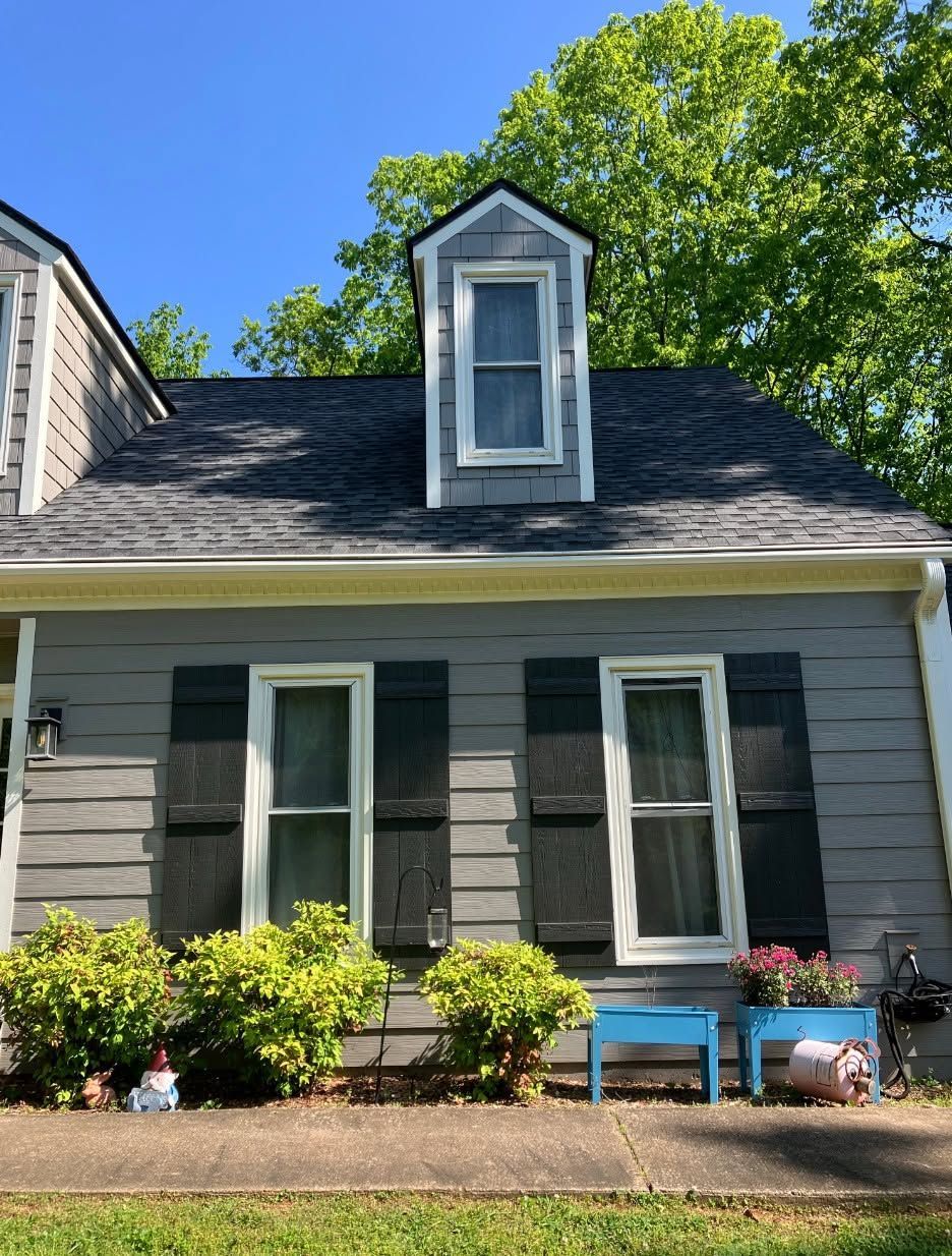 Gray house with white trim, dormer window, and flower boxes under a leafy green tree.