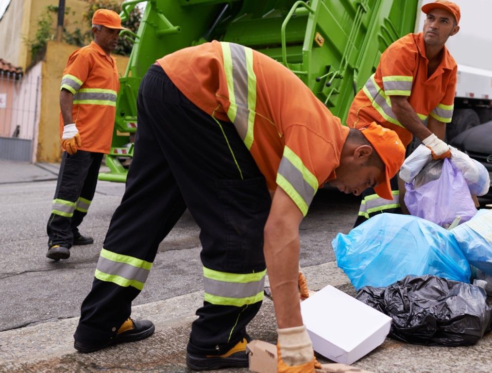 Workers in orange uniforms clean up trash beside a green garbage truck on a street.