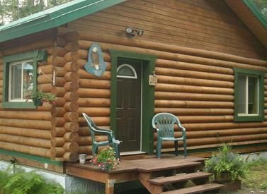 Log cabin with green trim and a small porch with two green chairs.