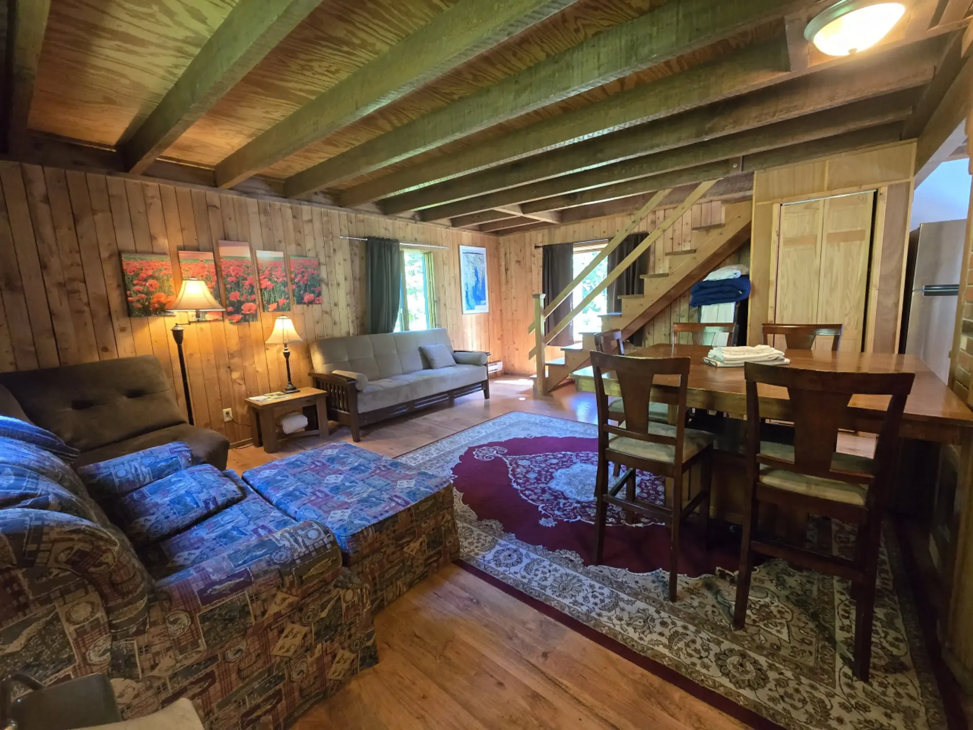 Cozy cabin interior with wood paneling, sectional sofa, futon, dining table, stairs, and rug.