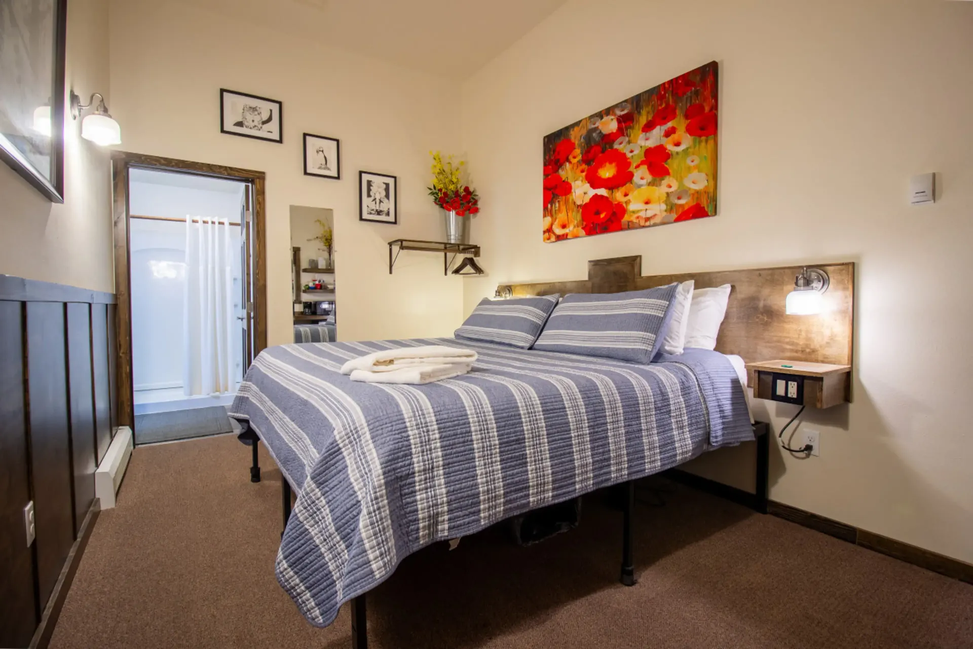 Cozy bedroom with bed, art, and an open bathroom door. Brown carpet, cream walls, and wood accents.