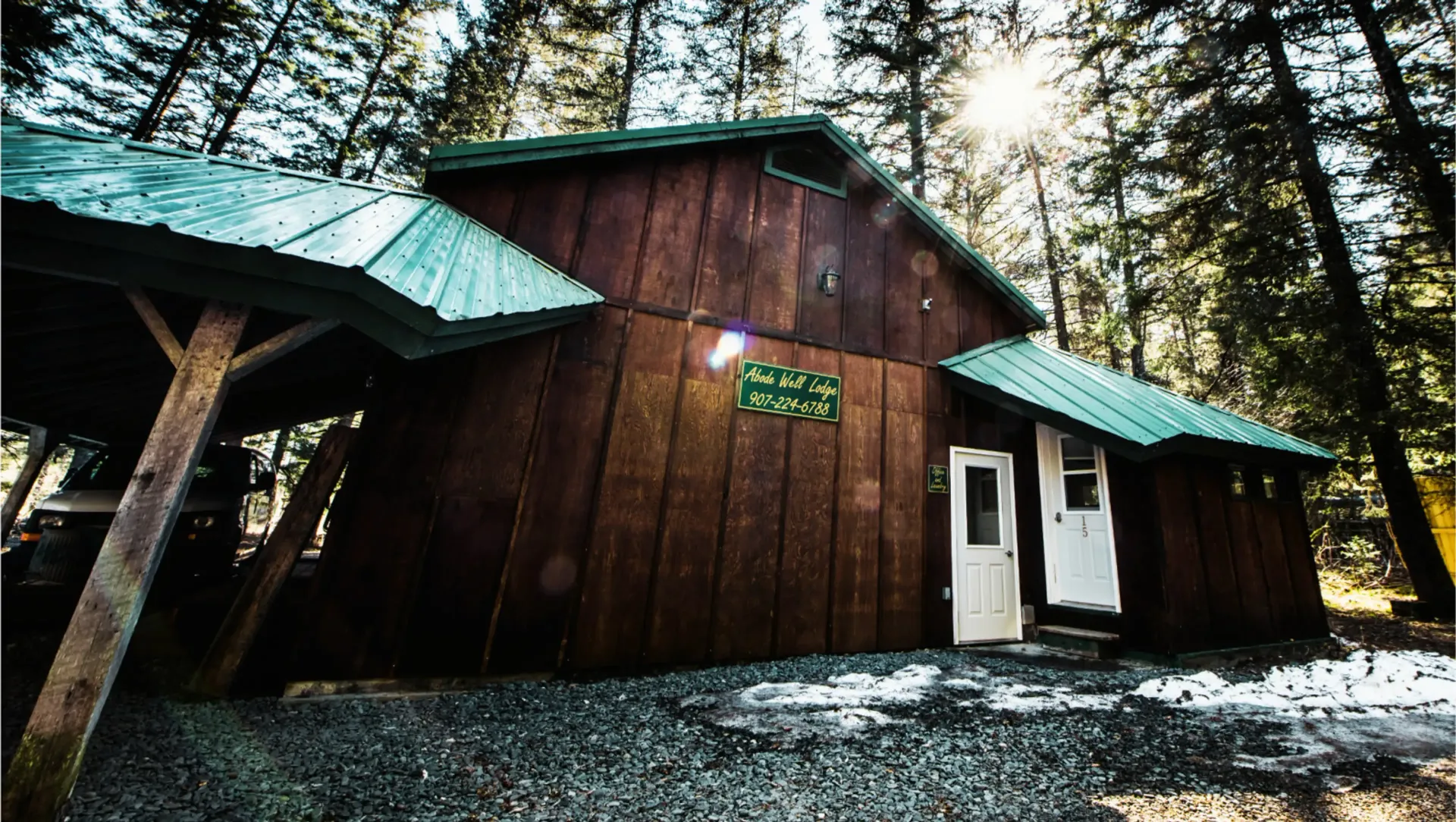 Wooden cabin with green roof and car shelter in a forest, sunlight shining.