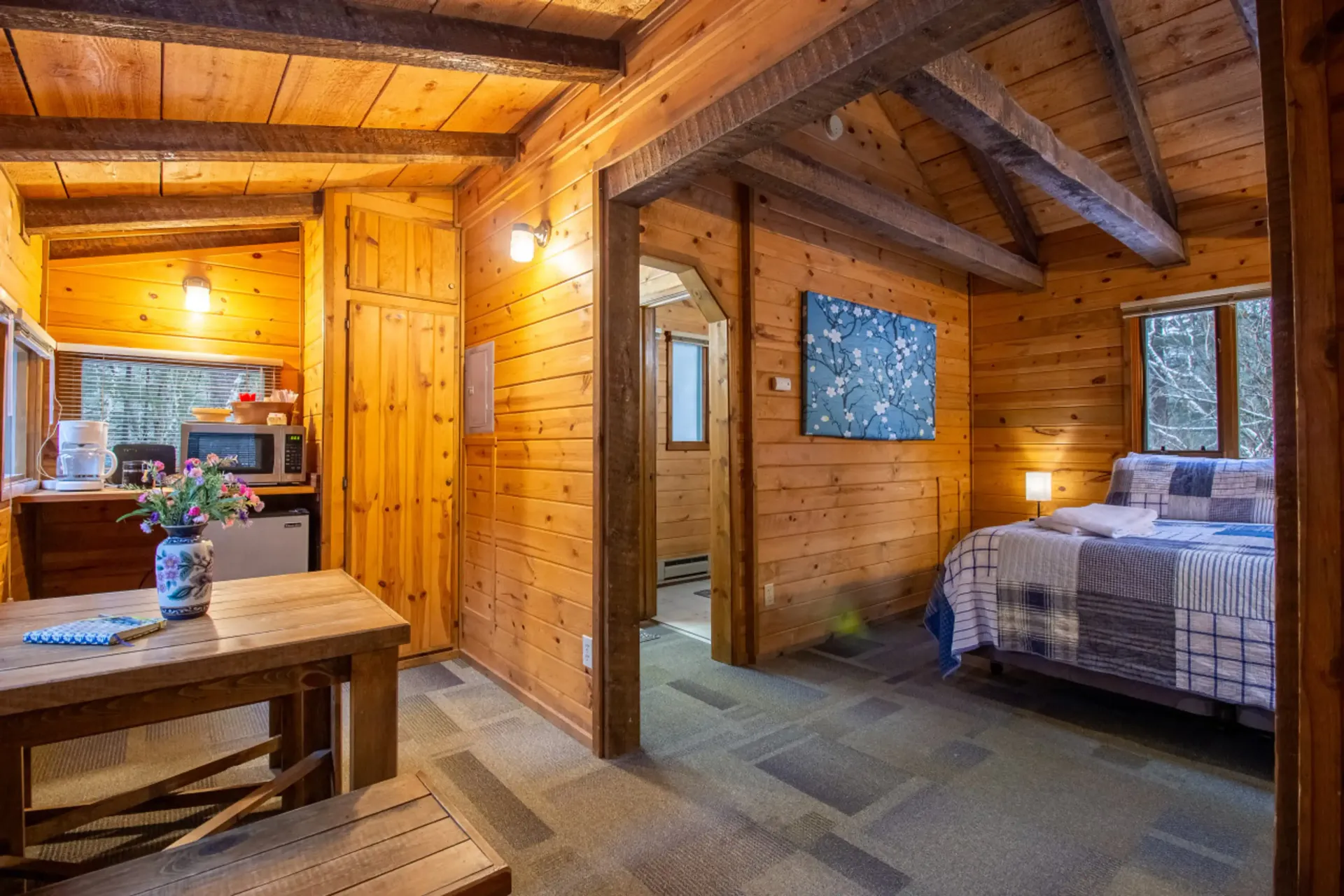 Cozy wood-paneled cabin interior with a bed, table, small kitchen, and an entryway to a bathroom.