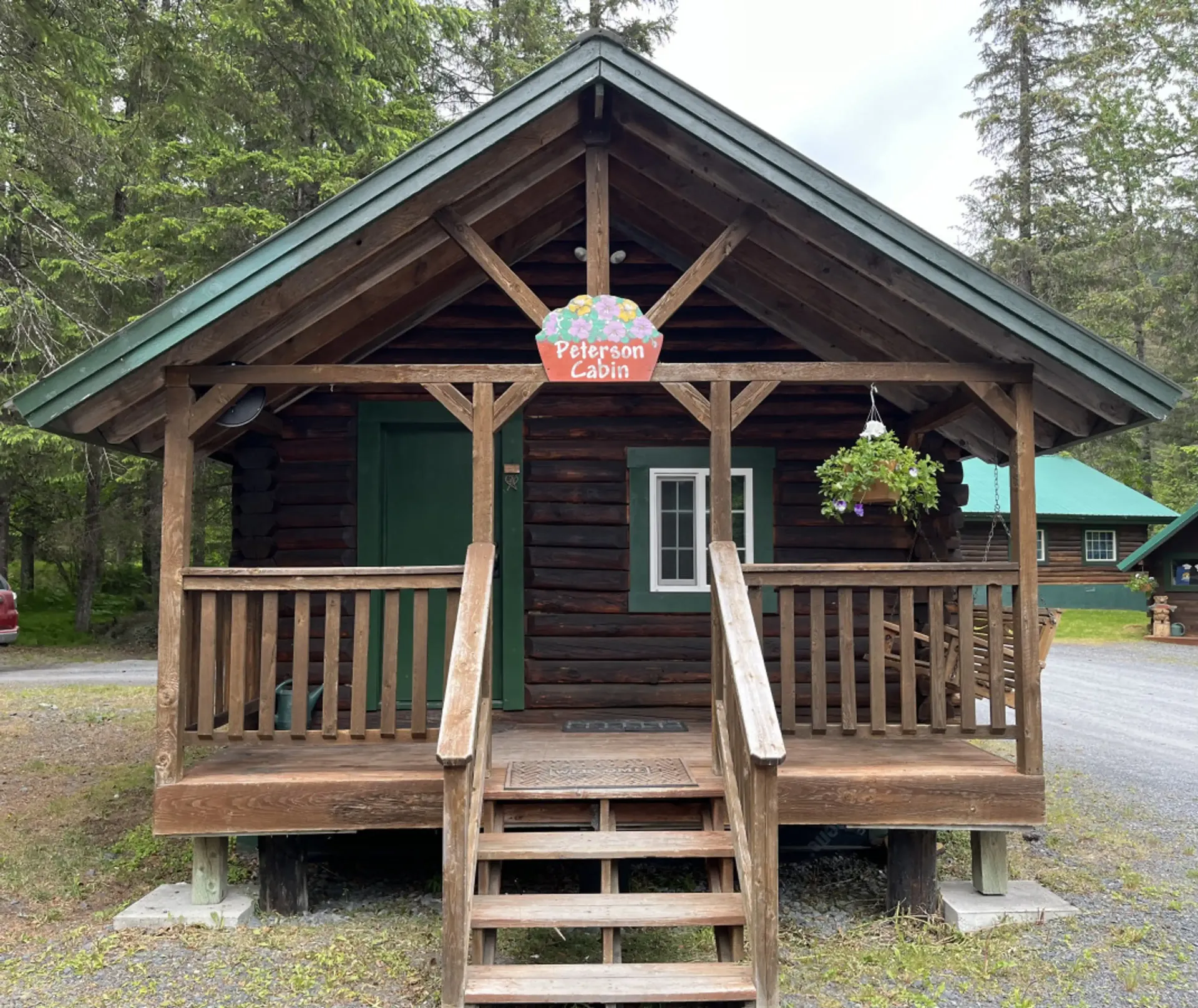 Log cabin with a porch and steps; green door, sign, and roof; flower basket; in a wooded setting.
