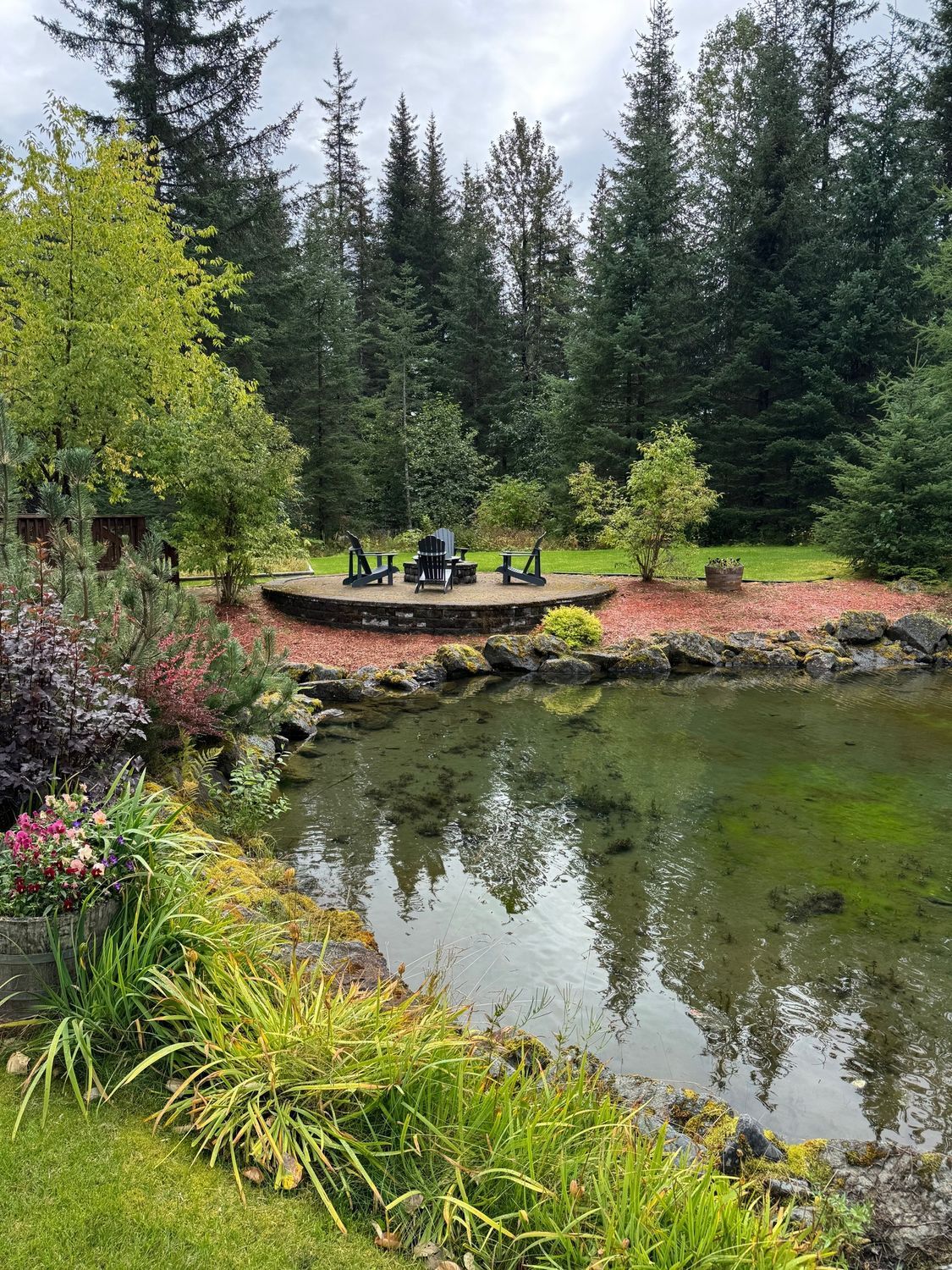 Pond in a lush garden with a small circular platform and chairs in front of a forest under a cloudy sky.