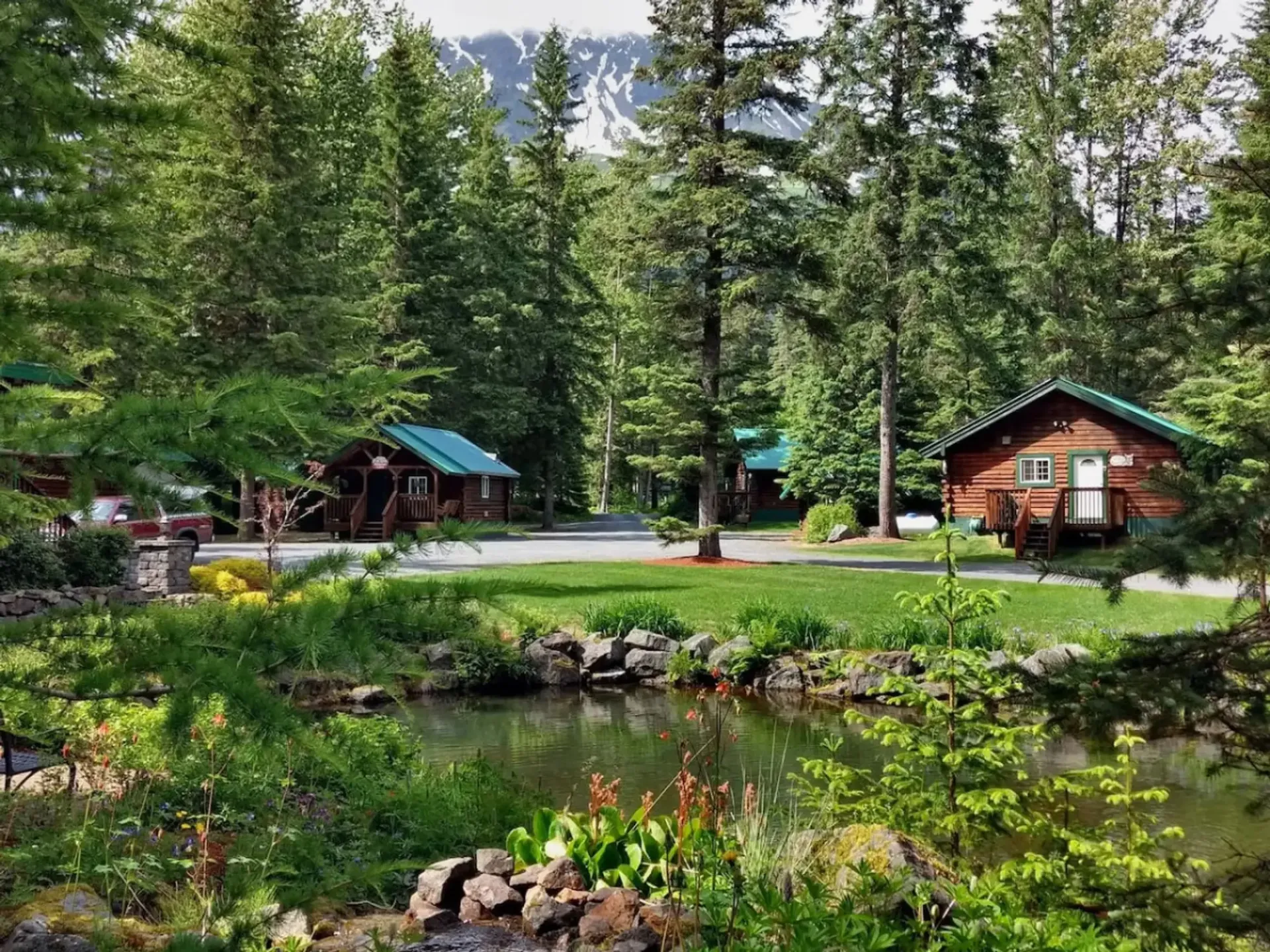 Cabins nestled in a forest with a pond in the foreground and a mountain in the distance.