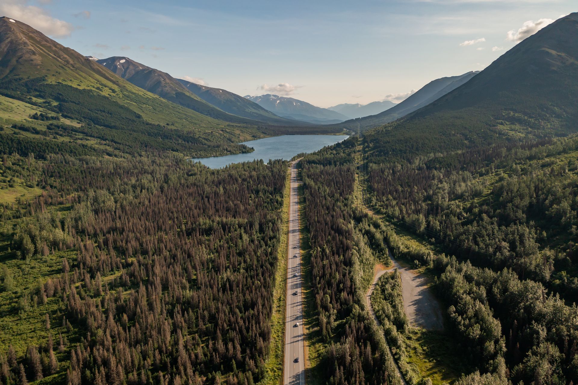 A road winds through a forest toward a lake, flanked by mountains under a blue sky.
