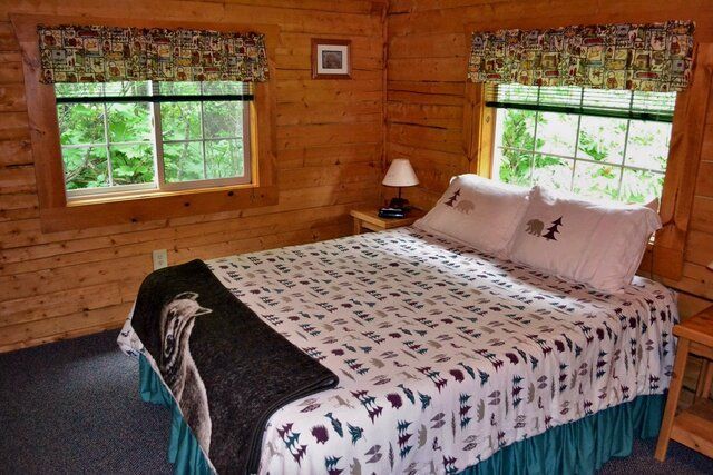 Cozy bedroom in a log cabin with a bed, windows, and nature views. Wooden walls, patterned bedding.