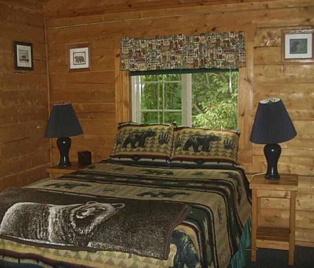 Bedroom with log walls, a bed with bear bedding, and two lamps. Window with green foliage.
