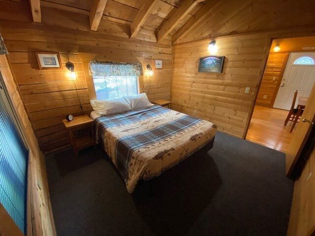 Bedroom with wood-paneled walls, a bed with a plaid comforter, and a doorway leading to another room.