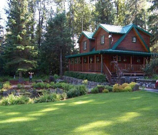 Log cabin with green roof in a forest setting, featuring a wraparound porch and landscaped yard.