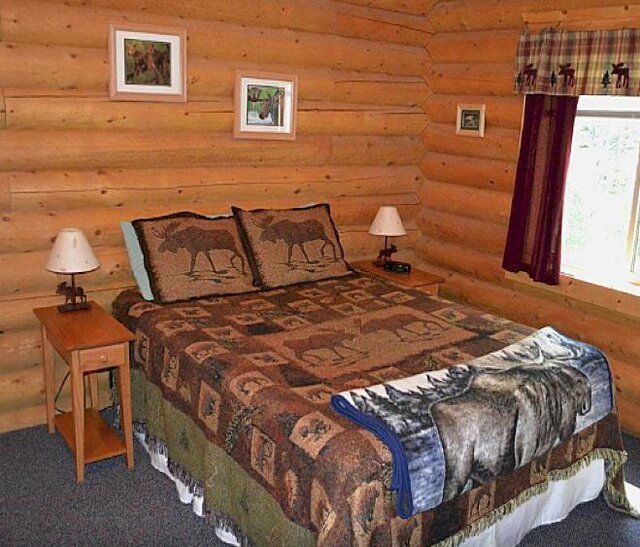 Cozy cabin bedroom with a bed featuring moose decor. Log walls, two bedside tables, and a window with curtains.