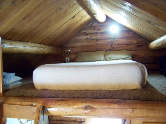 A loft bedroom with a bed, built into a cabin, beneath wooden logs.