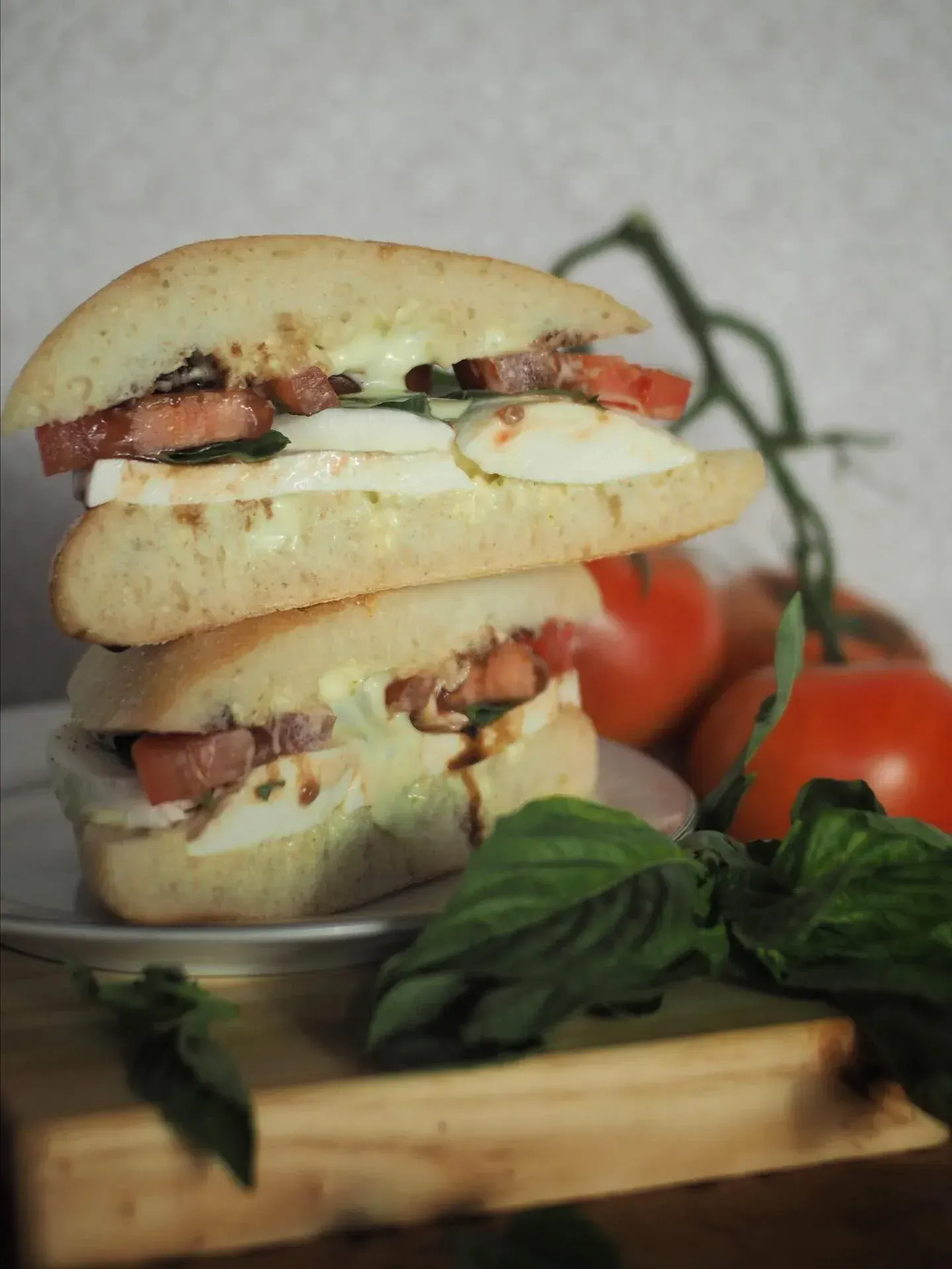 Two stacked sandwiches with tomato, mozzarella, and basil; tomatoes and basil leaves in background.