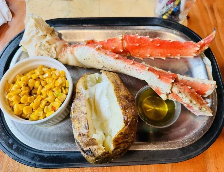 King crab leg, baked potato, corn, and butter on a tray.