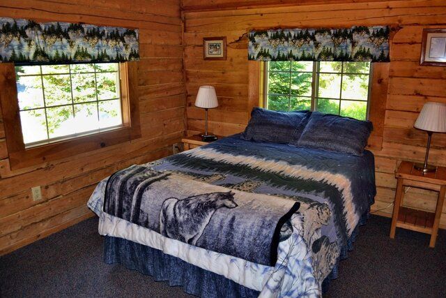 Bedroom in a log cabin with a bed featuring a wolf blanket, two windows, and wood paneling.