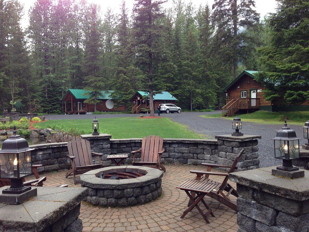 Stone patio with fire pit and seating, overlooking cabins in a wooded area.