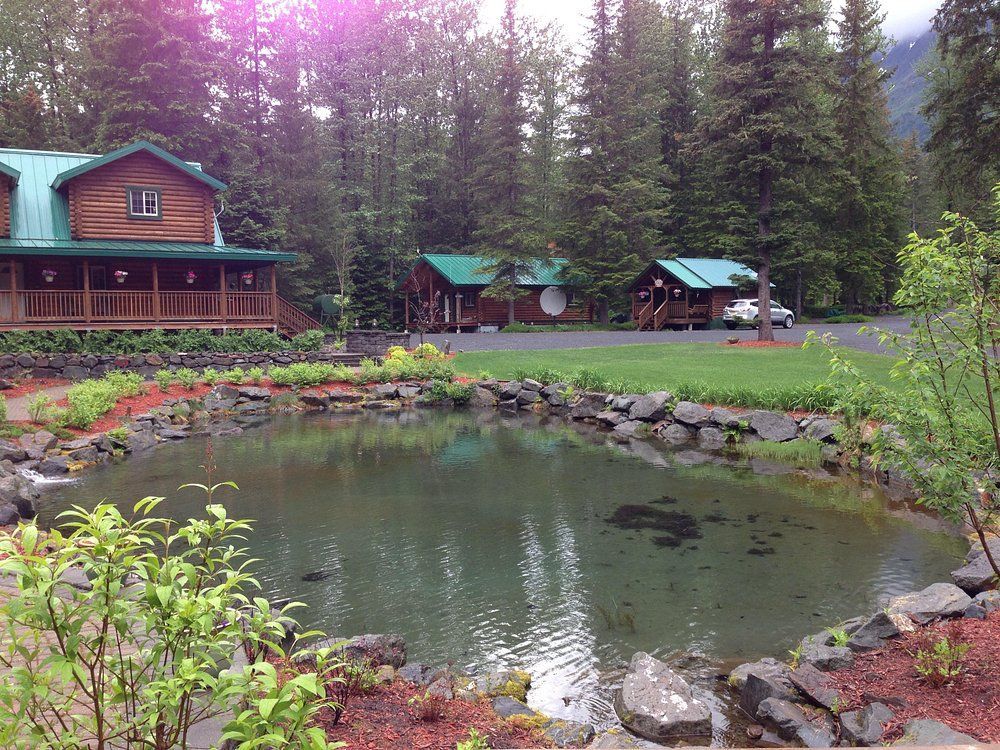 Lodge with green roof overlooking a pond. Cabins and trees in the background, cloudy sky.
