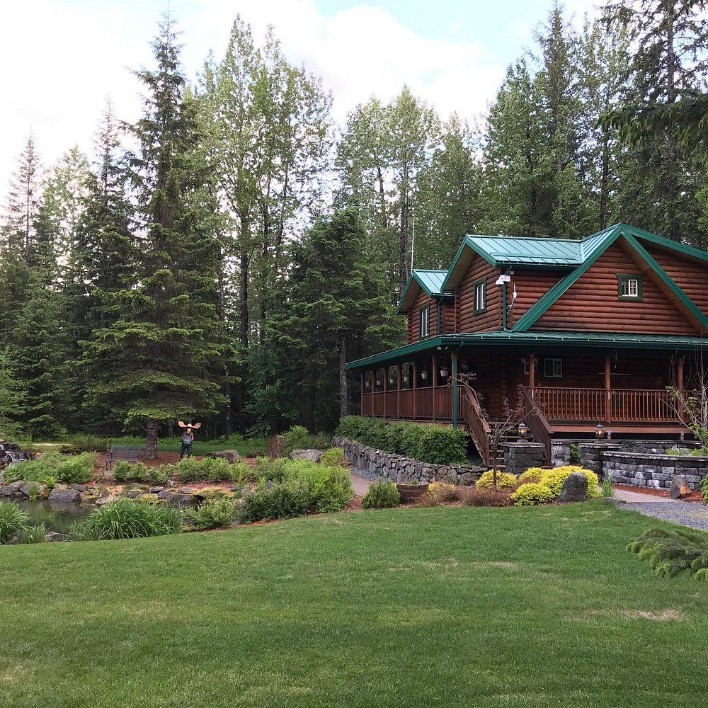 Log cabin nestled in lush green forest. A person stands on a lawn with arms raised.