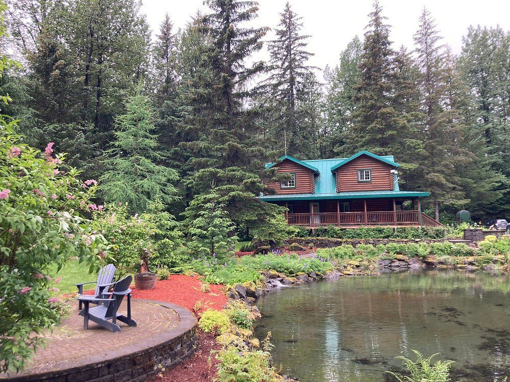Cabin with green roof and wrap-around porch sits beside a pond surrounded by trees and flowers.