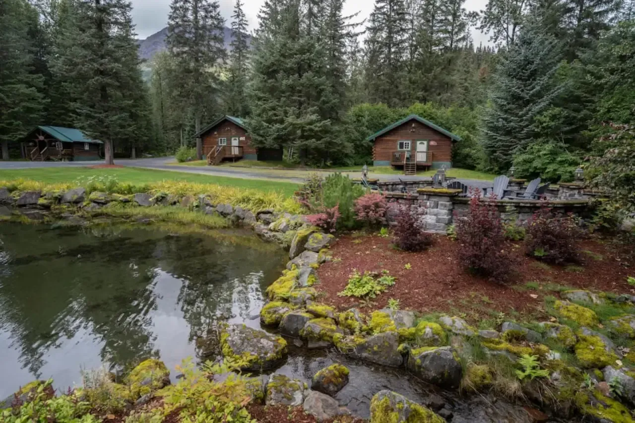 Cabins by a pond in a forest setting. Green trees surround small buildings and a rocky pond with a waterfall.