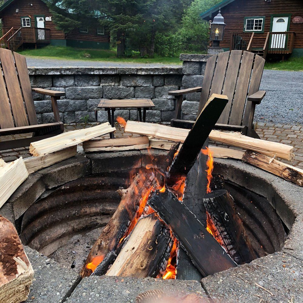 Fire pit with burning logs, Adirondack chairs, cabins in background.
