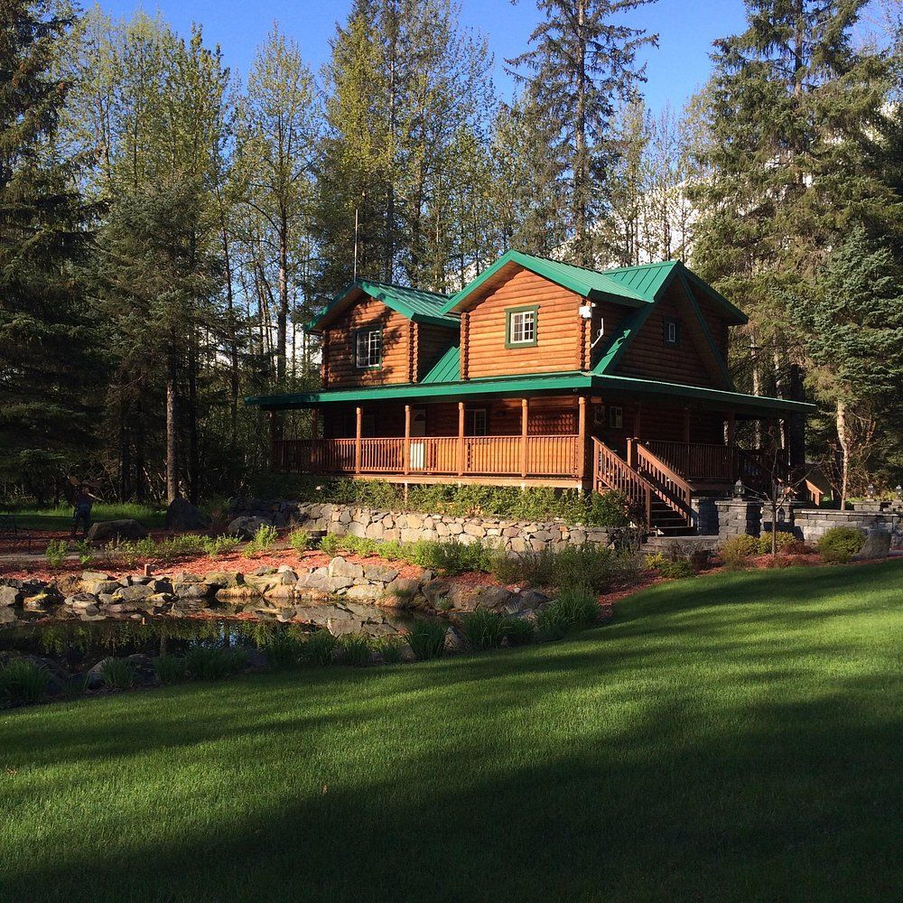 Log cabin with green roof surrounded by trees and a grassy lawn.