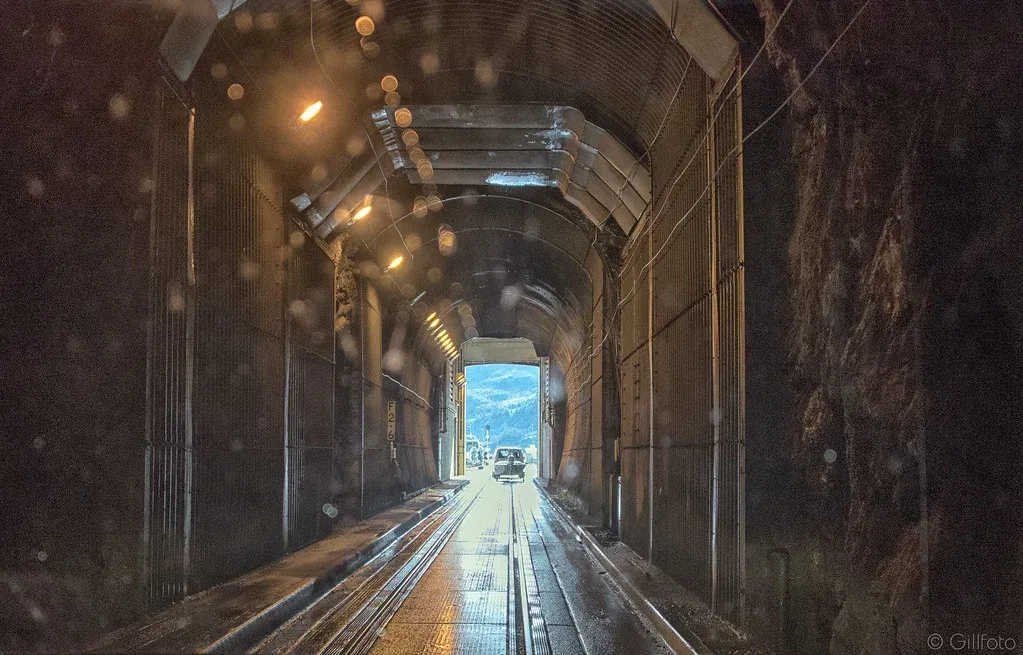 Dark tunnel interior with rain falling. A vehicle is visible at the tunnel's exit, revealing daylight.