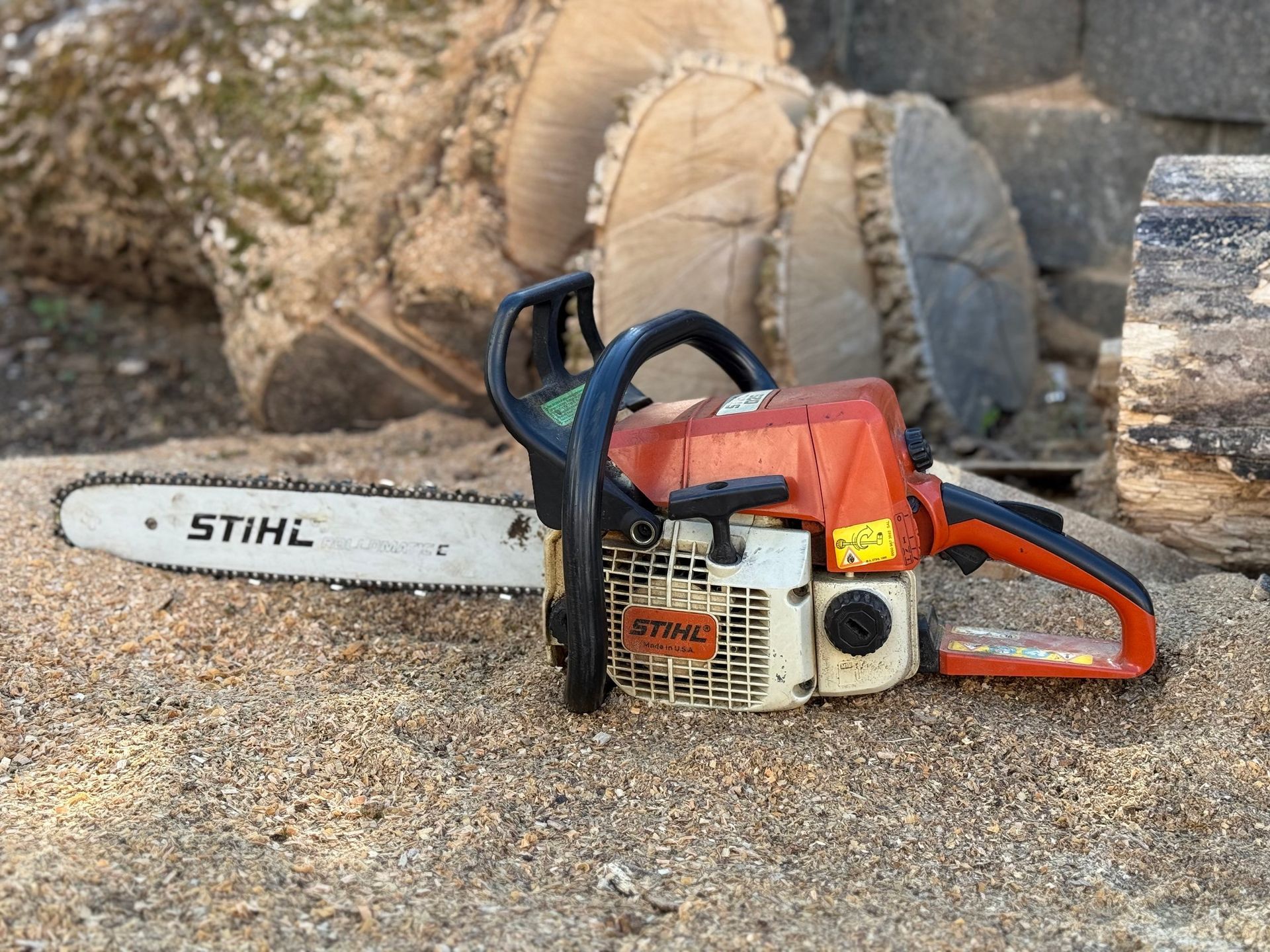 A Stihl chainsaw with an orange and white body lies on wood chips, with cut logs in the background.