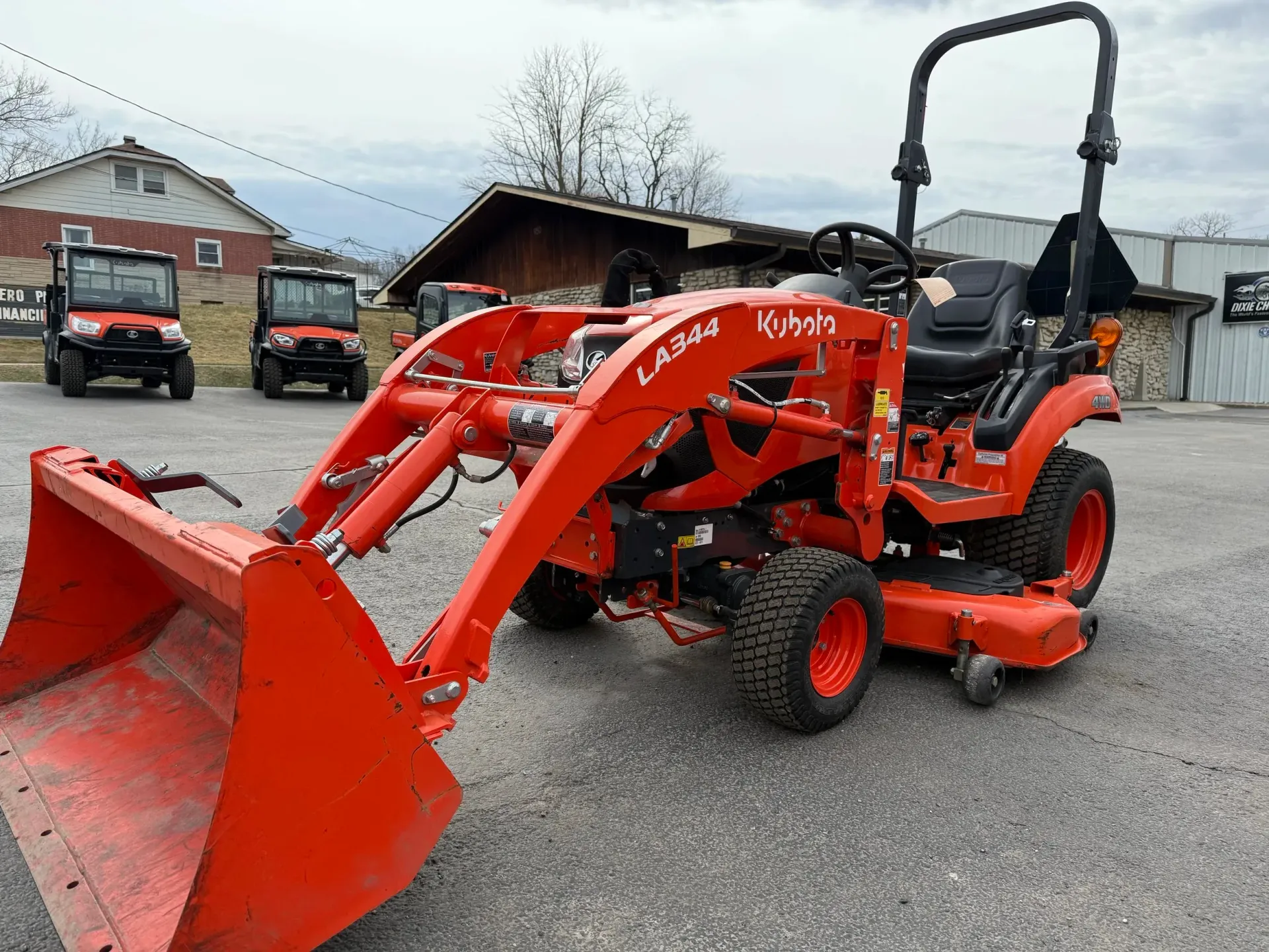 Orange Kubota tractor with a front-end loader on display, parked outside. Other vehicles visible in background.